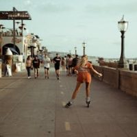 Woman roller skating on a sunny beachside promenade, surrounded by pedestrians and classic architecture.